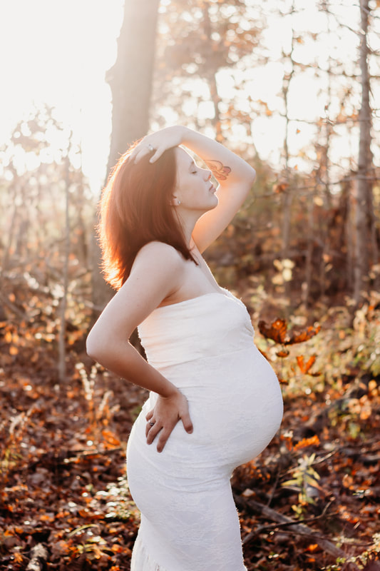 Maternity photo. woman standing with leg slightly bent, one hand in hair, eye closed, head tilted up. Warm sunshine shining down from behind her. Park in Fort Wayne, Indiana